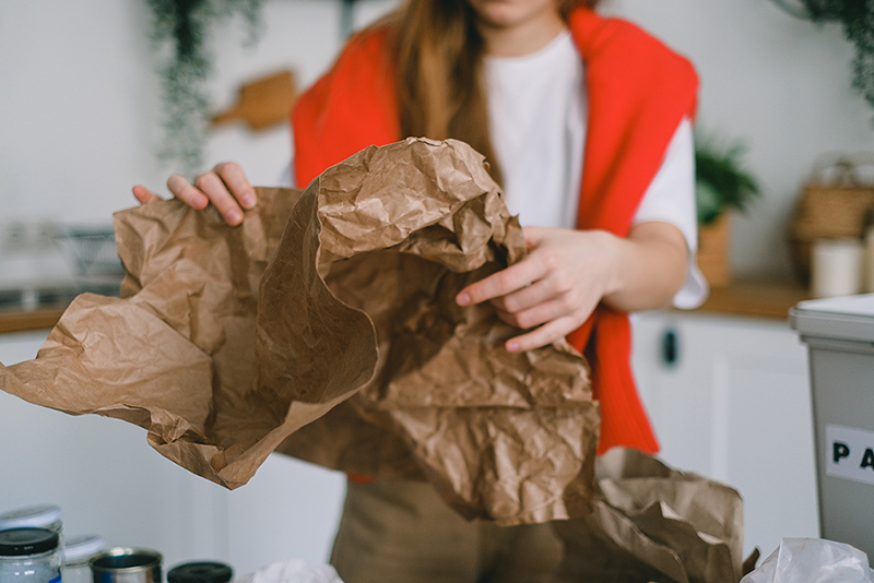 Person in a red cardigan holding crumpled brown paper in a kitchen setting, symbolizing eco-friendly practices like composting or repurposing paper waste.